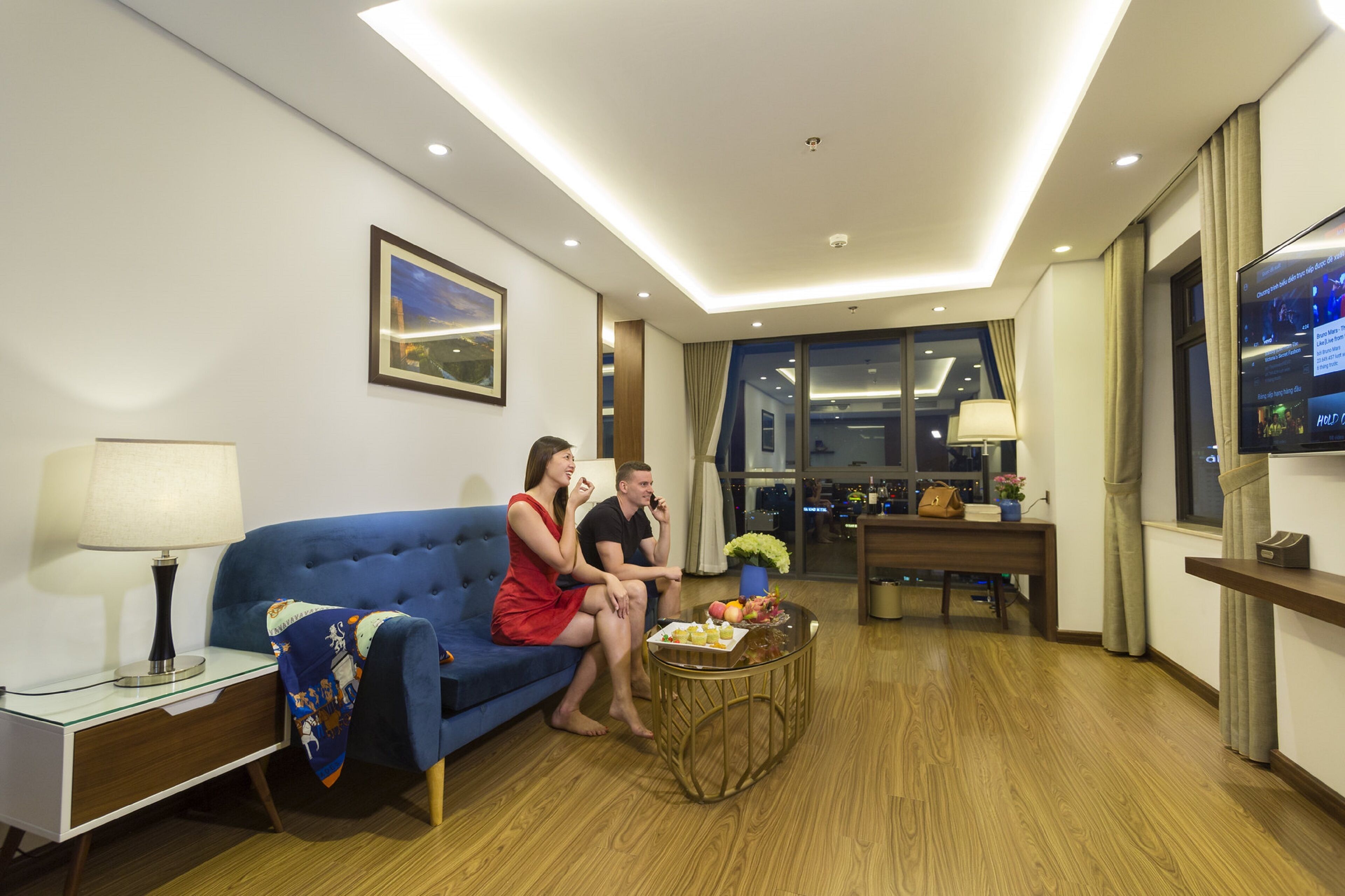 Una habitación de hotel moderna con un sofá azul, una mesita redonda con fruta, un televisor y un escritorio con una lámpara.