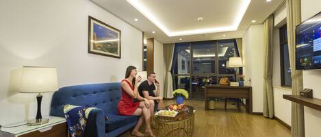 Una habitación de hotel moderna con un sofá azul, una mesita redonda con fruta, un televisor y un escritorio con una lámpara.