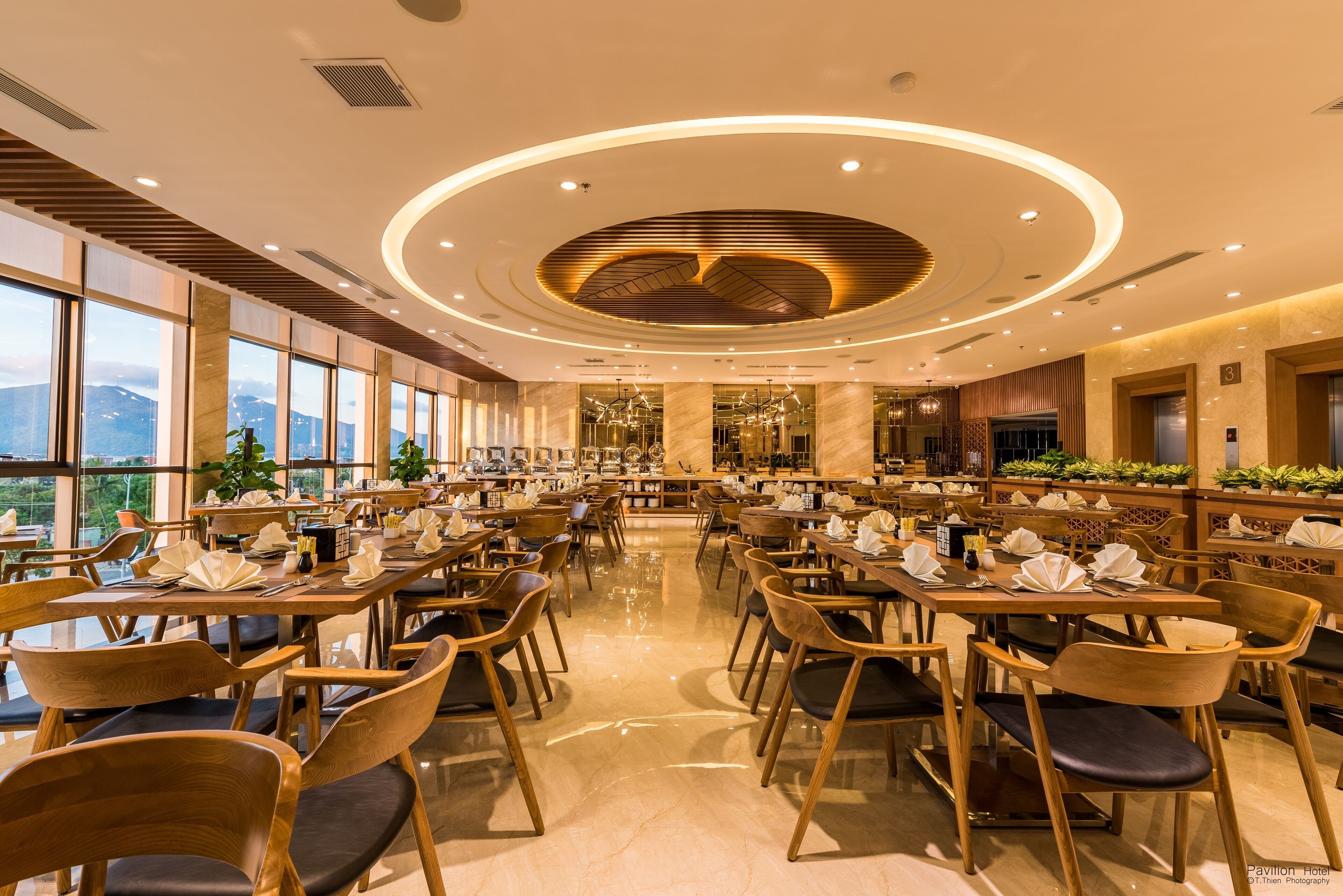 Un restaurante amplio con varias mesas y sillas, grandes ventanales que ofrecen vistas a las montañas y un diseño de techo moderno con iluminación circular.