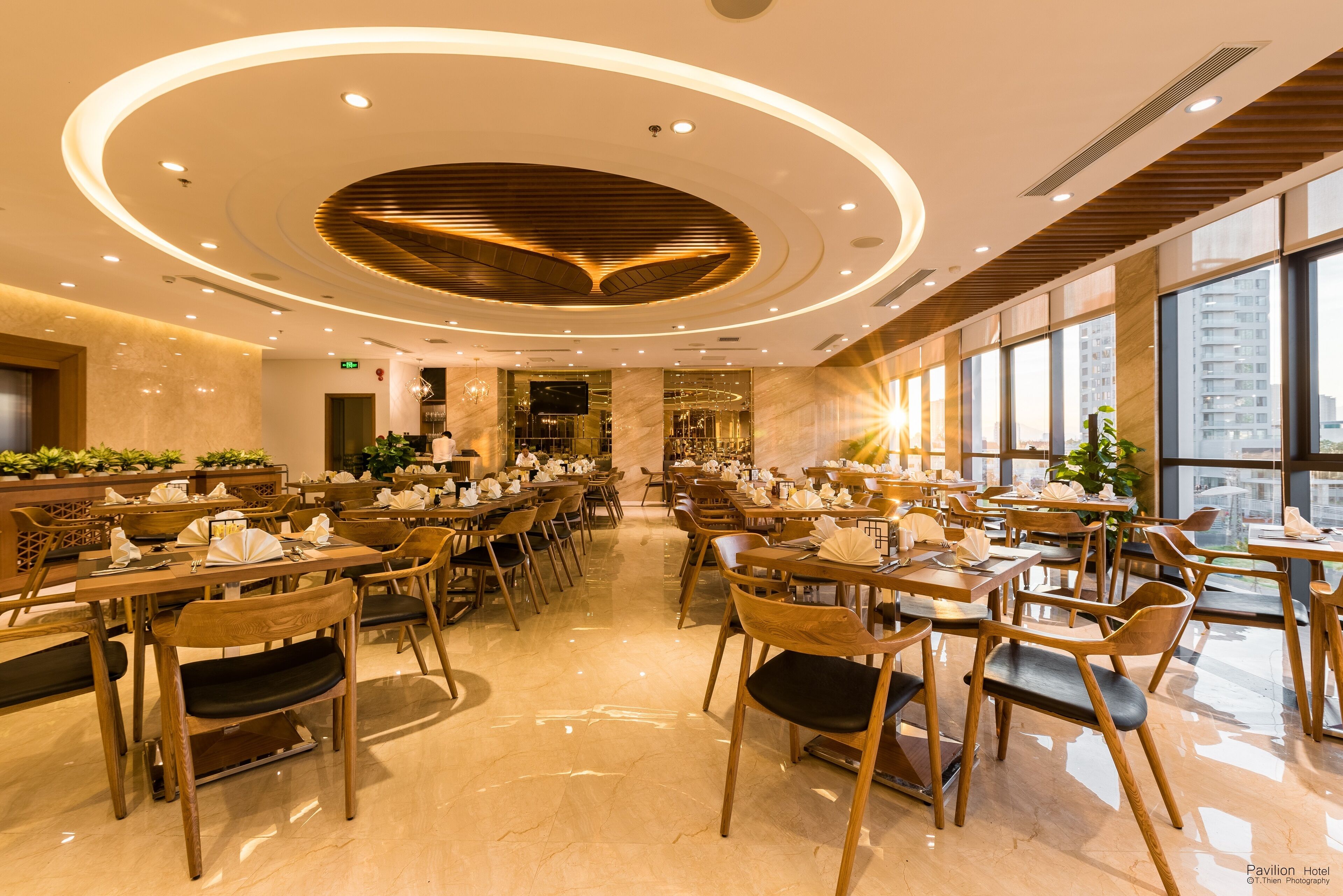 A spacious hotel dining area with multiple tables and chairs, large windows offering views of the city, and a modern ceiling design.