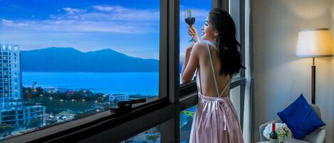 Una habitación con un ventanal grande con vistas a una ciudad y un cuerpo de agua, una mesa con una botella de vino y una lámpara.