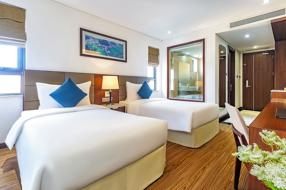 Quarto de hotel com duas camas, uma escrivaninha e um banheiro visível por uma porta de vidro.