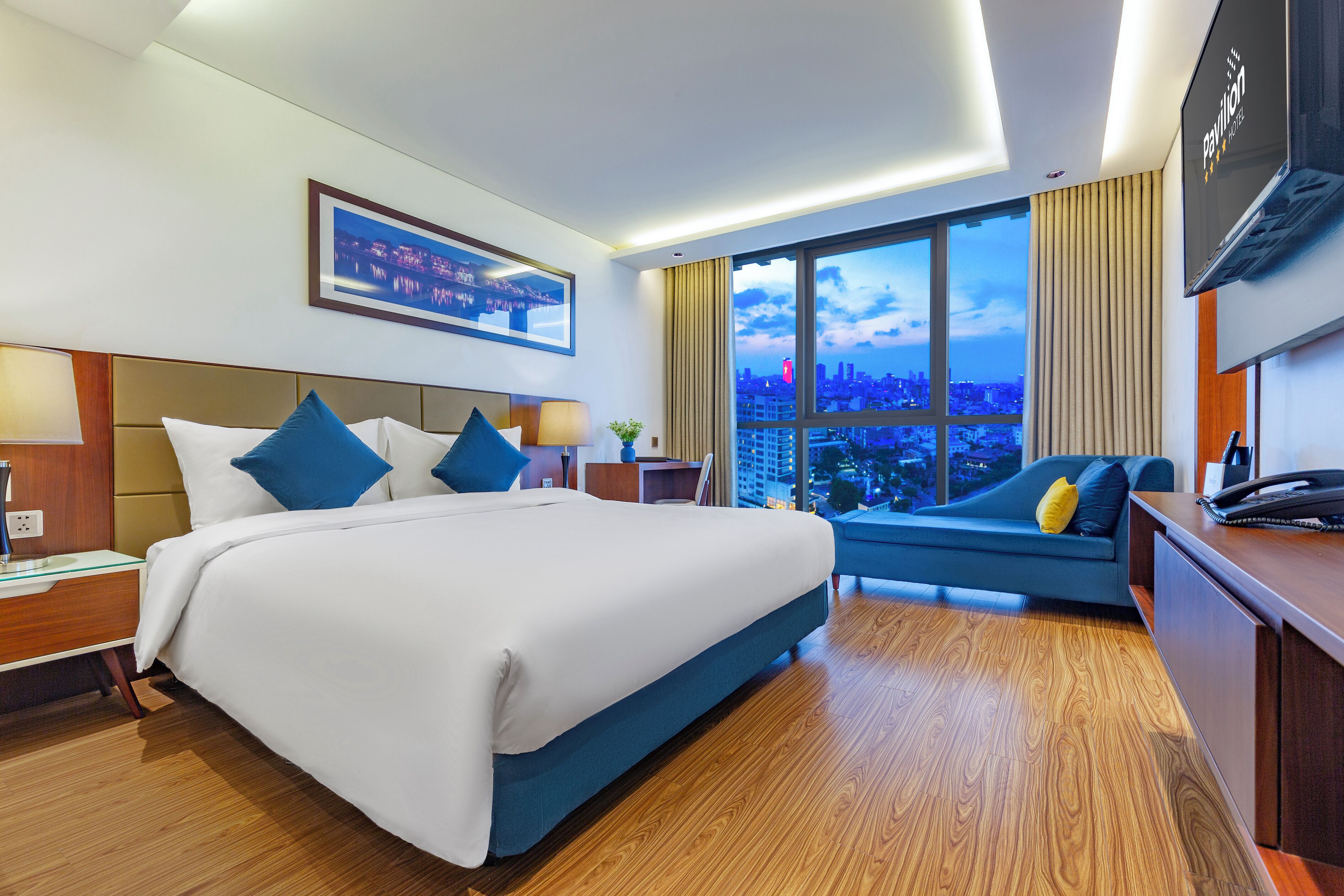 A hotel room with a large bed, a desk, a chair, a TV, and a view of the city.