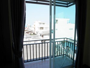 A balcony with a glass door and a metal railing, overlooking a residential area with white buildings. - Baan Yooyen Hua Hin 57 (Hua Hin)