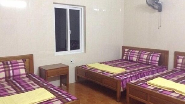 Een kamer met drie houten bedden, elk voorzien van een paars-wit geruit sprei en een gele deken. Er is een raam met witte kaders en een wandgemonteerde airconditioning.
