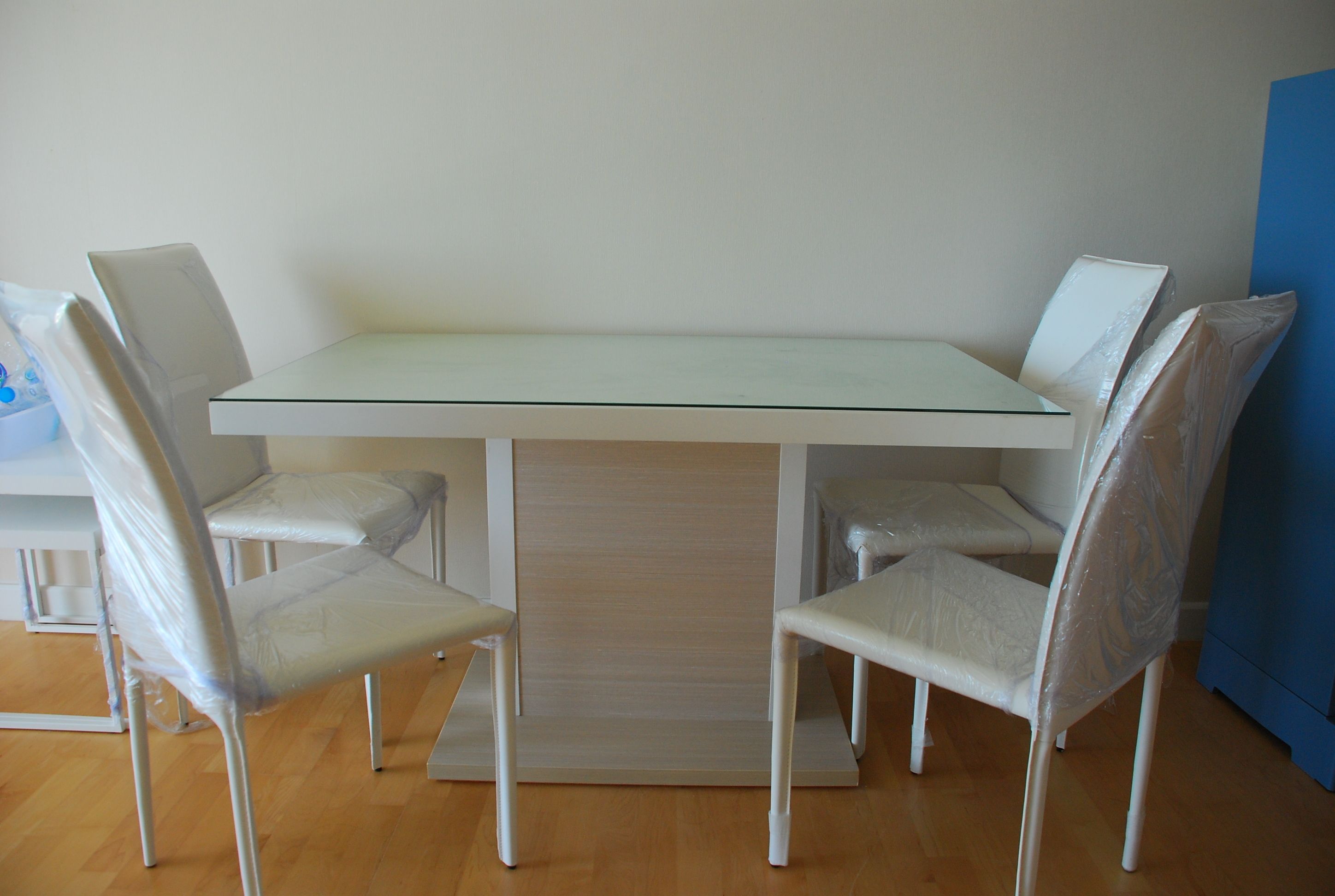 A dining table with four chairs, a light-colored floor, and a plain wall.
