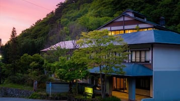A two-story building with a sloped roof, surrounded by lush greenery and a hillside.
