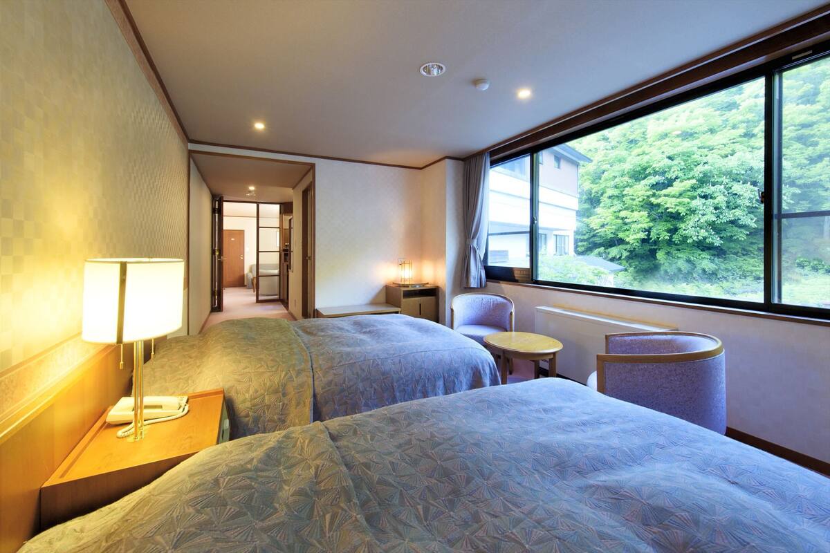 A hotel room with two beds, a small table, a chair, a lamp, and a window with a view of greenery outside.