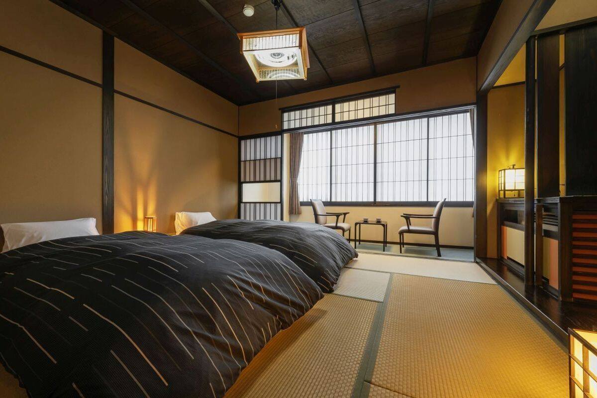 A traditional Japanese-style room with tatami flooring, a large bed with a dark striped bedspread, a small table, and a chair.