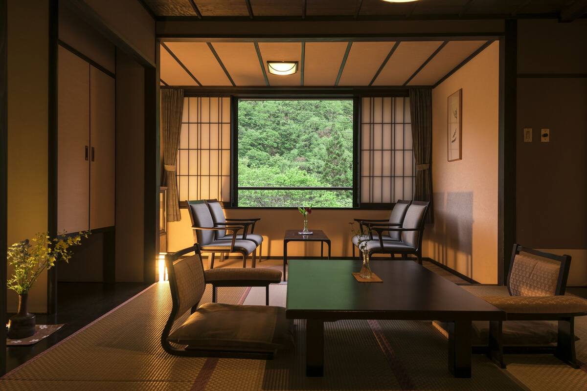 A traditional Japanese room with tatami flooring, a low table, and four chairs. A large window offers a view of a forest.