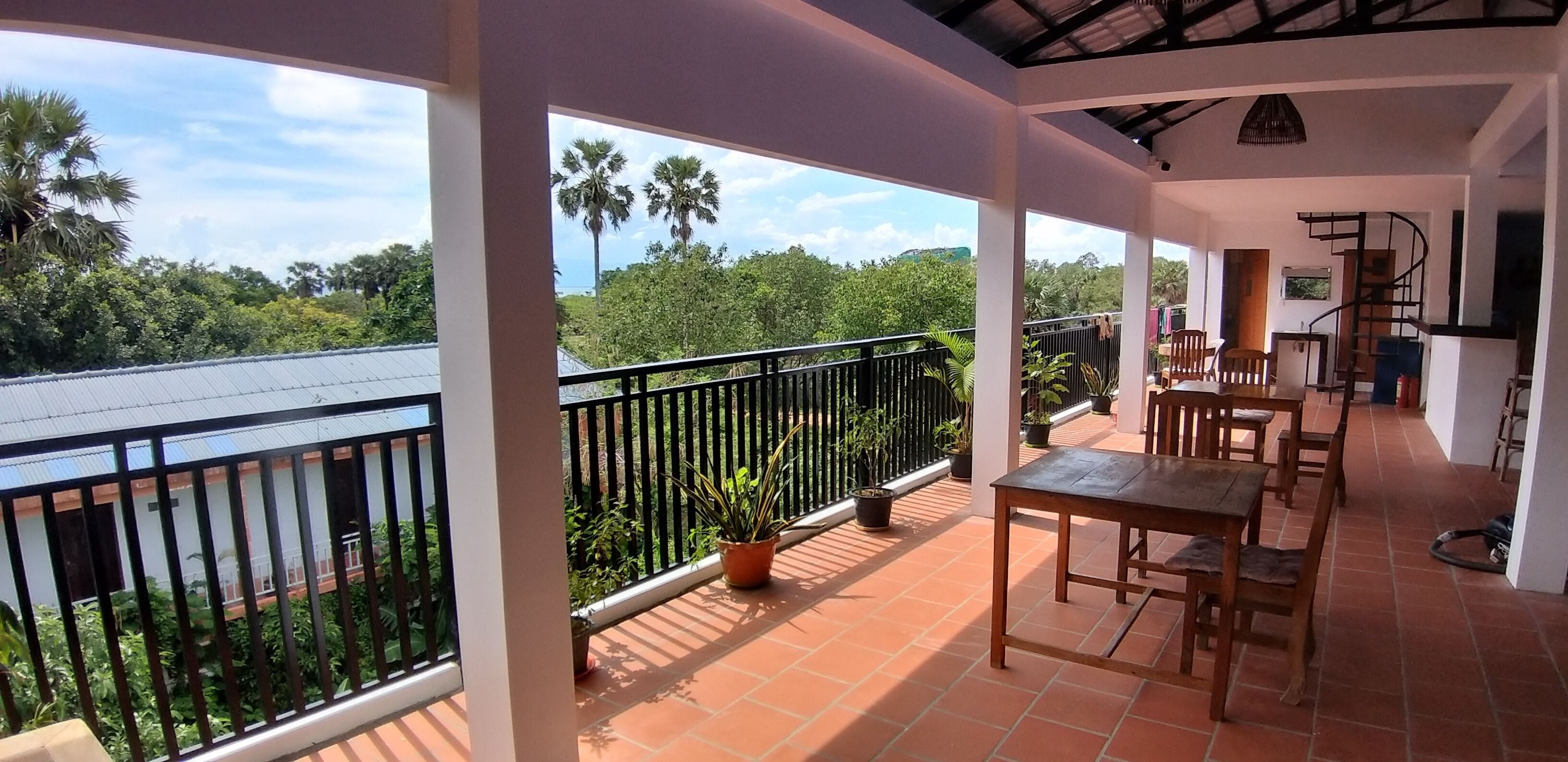 A covered balcony with a view of palm trees and a river. There are wooden tables and chairs, and potted plants.
