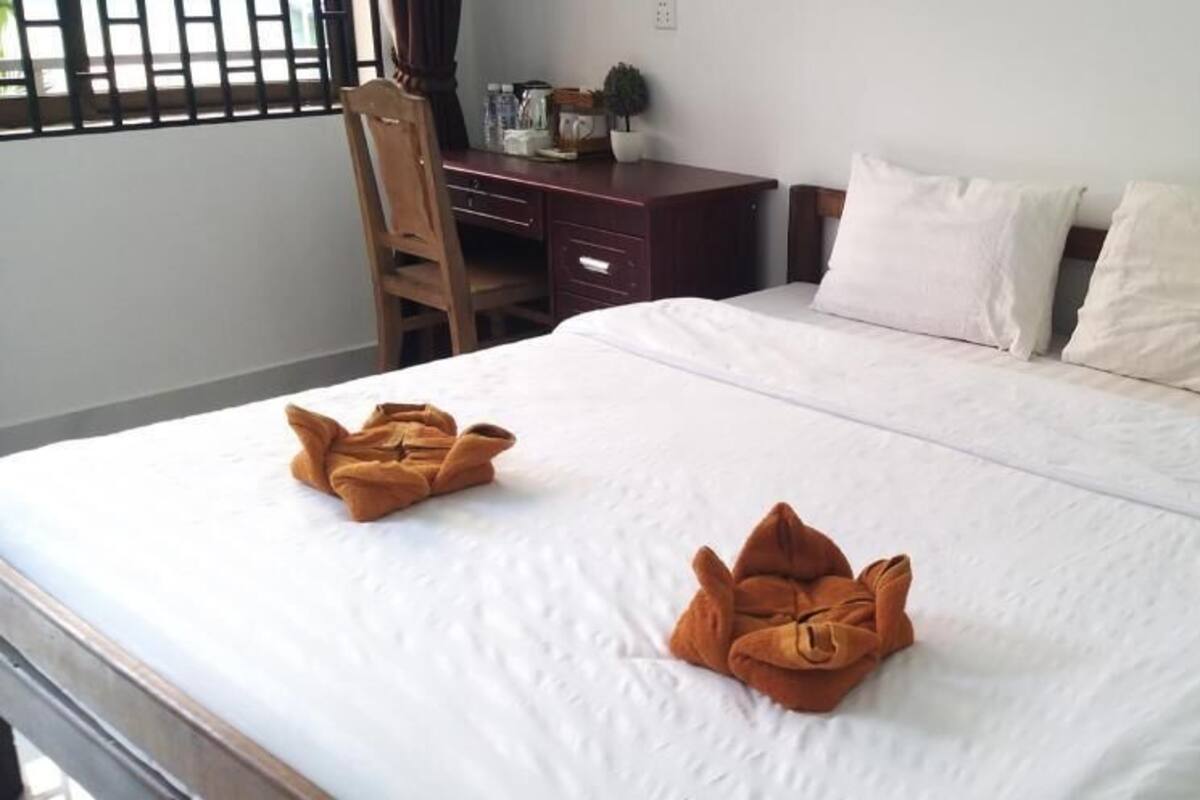 A hotel room with a bed, two pillows, a wooden nightstand, a desk, and a chair.
