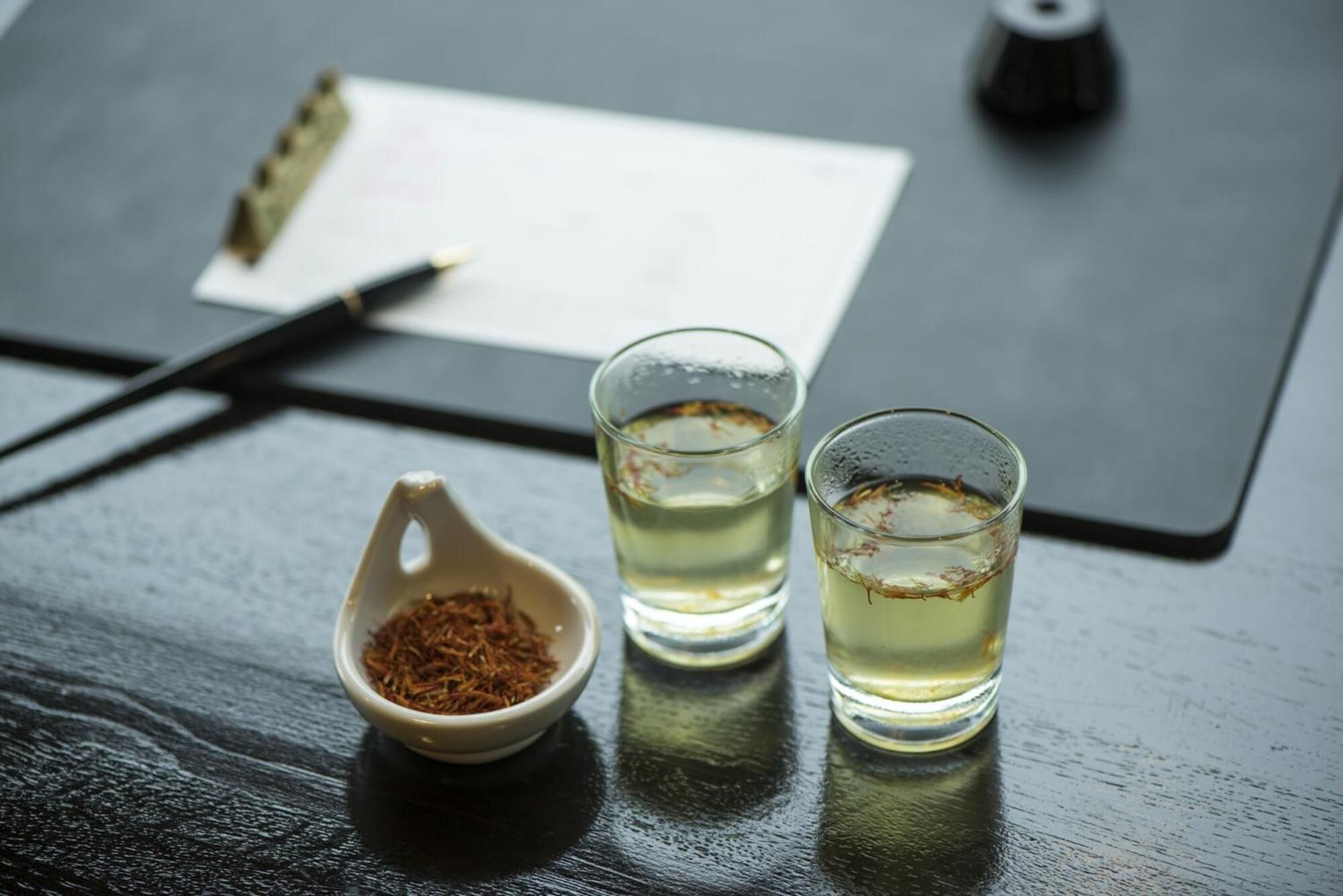 Uma mesa de madeira escura com uma chávena de cerâmica branca cheia de um líquido amarelado, dois copos de vidro transparente com líquido semelhante e uma pequena travessa de cerâmica branca com uma substância castanha.