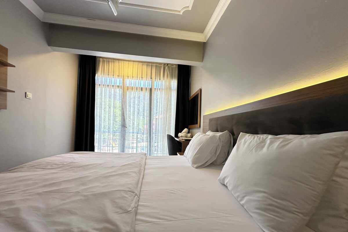 A hotel room with a large bed, a bedside table with a lamp, and a window with curtains.