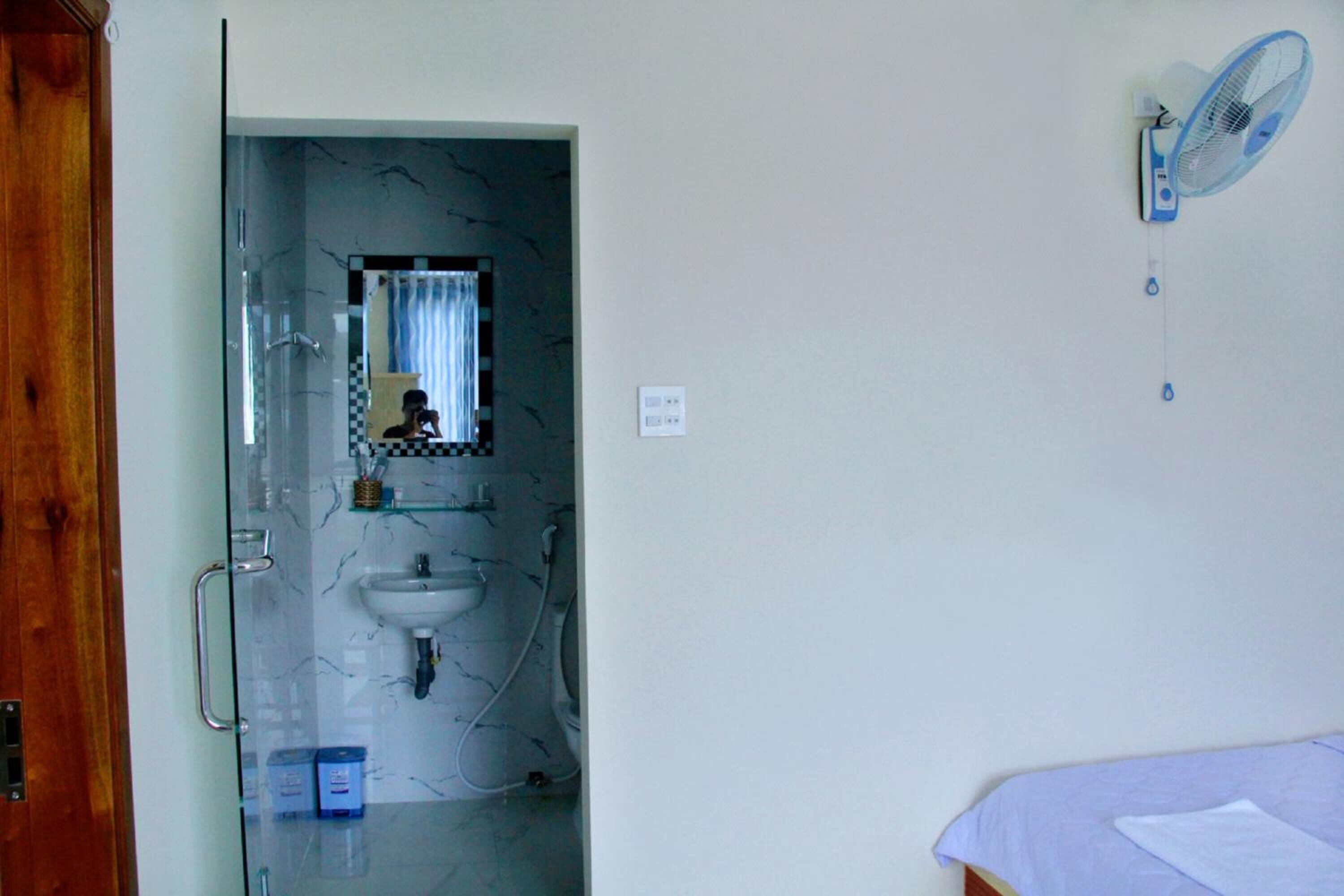 A bathroom with a glass shower door, a sink, and a mirror. There is a blue fan on the wall and a bed with a white sheet.