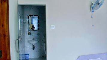 A bathroom with a glass shower door, a sink, and a mirror. There is a blue fan on the wall and a bed with a white sheet.