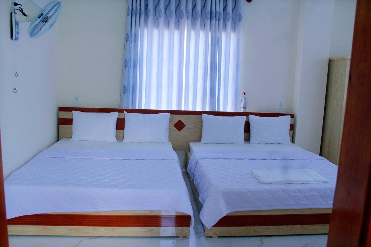 A room with two single beds, a wooden headboard, white bedding, a window with sheer curtains, and a wall-mounted fan.