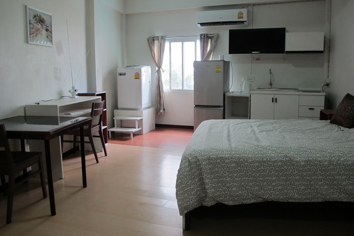 A bedroom with a bed, a desk, chairs, a refrigerator, a television, and a window with curtains.