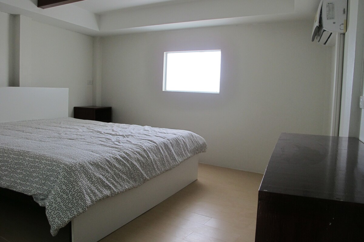 A bedroom with a bed, a small bedside table, a window, and a dark piece of furniture.