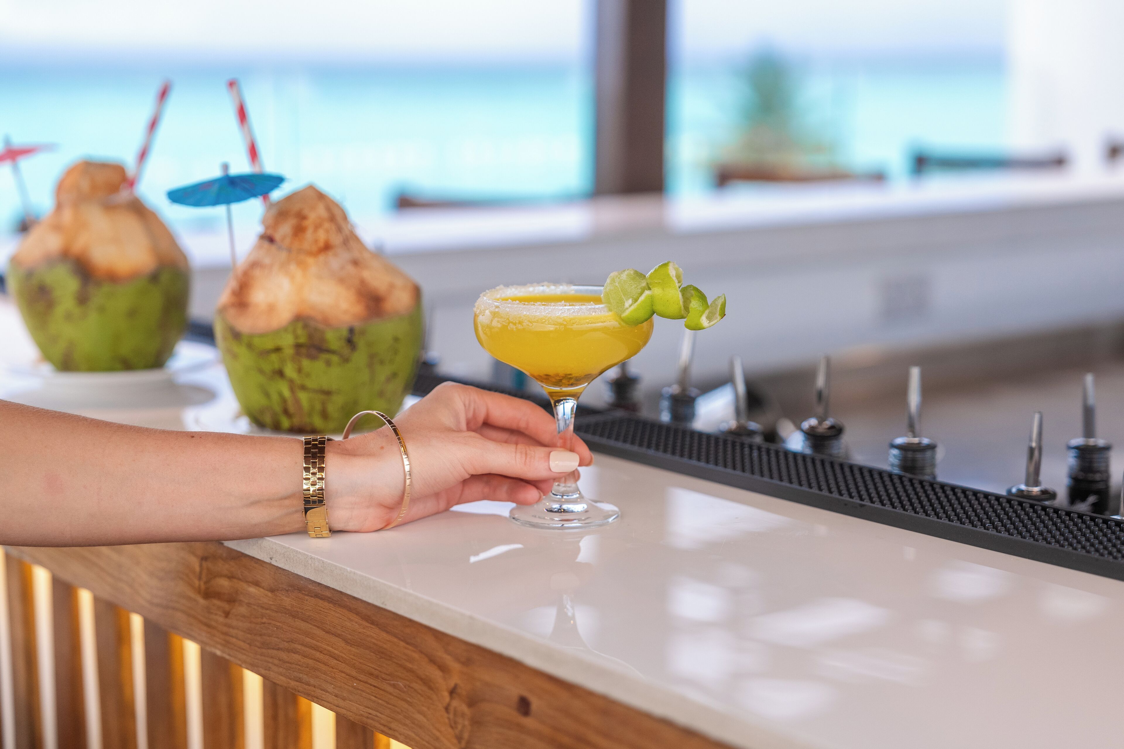 A bar counter with a drink, a coconut, and a person holding a glass.
