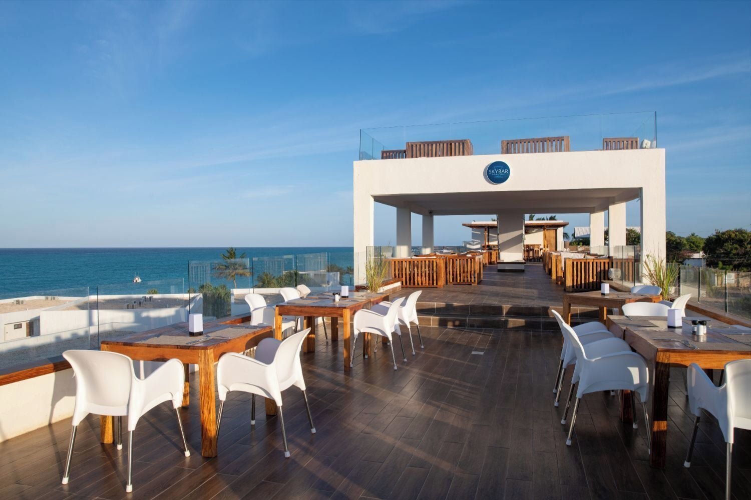 Un espace de restauration aménagé sur le toit, offrant une vue sur la mer, avec des tables en bois et des chaises blanches, ainsi qu’une enseigne « Skybar ».