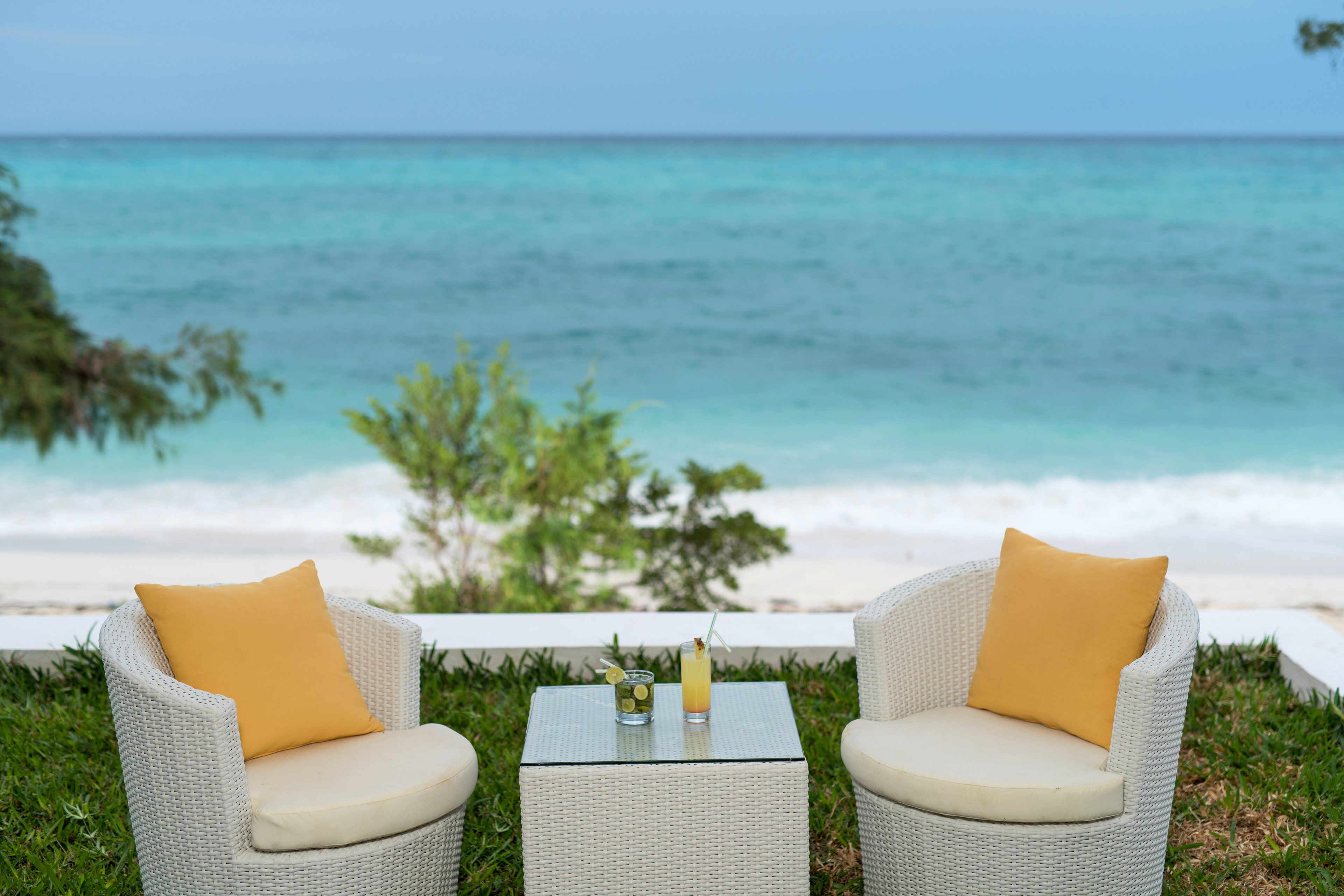 Deux fauteuils en osier avec des coussins jaunes et une petite table avec des boissons, le tout placé devant une vue sur la plage.