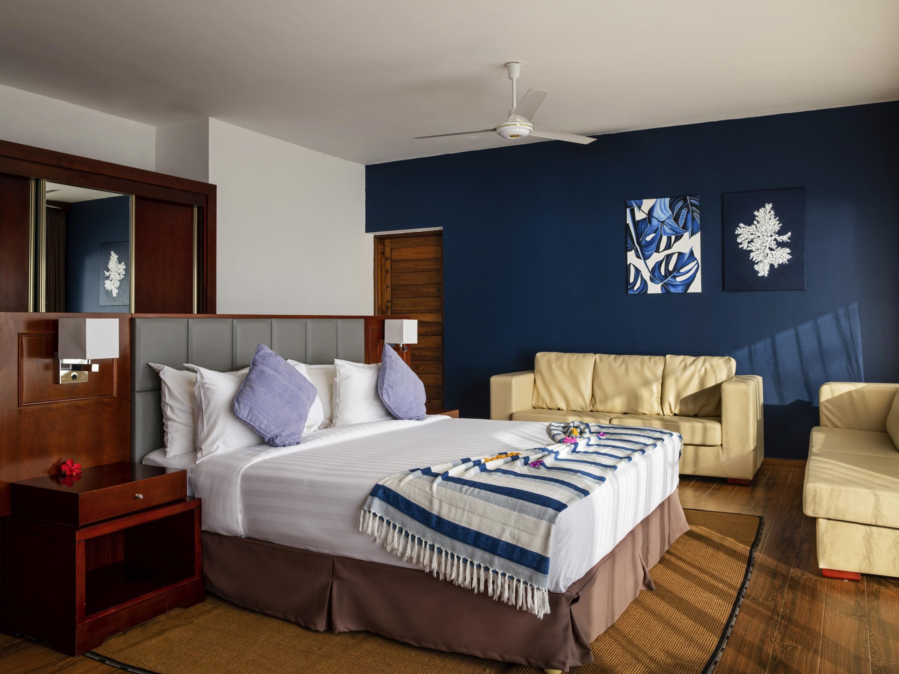 A hotel room with a bed, a sofa, a nightstand, a ceiling fan, and artwork on the walls.