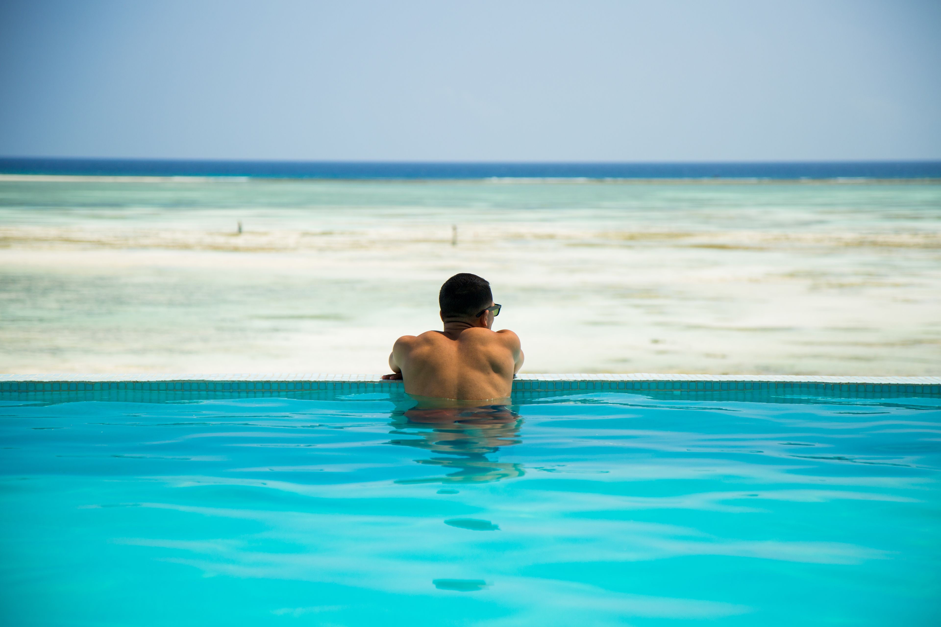 A man sitting in a swimming pool with a view of the ocean and a sandy beach.