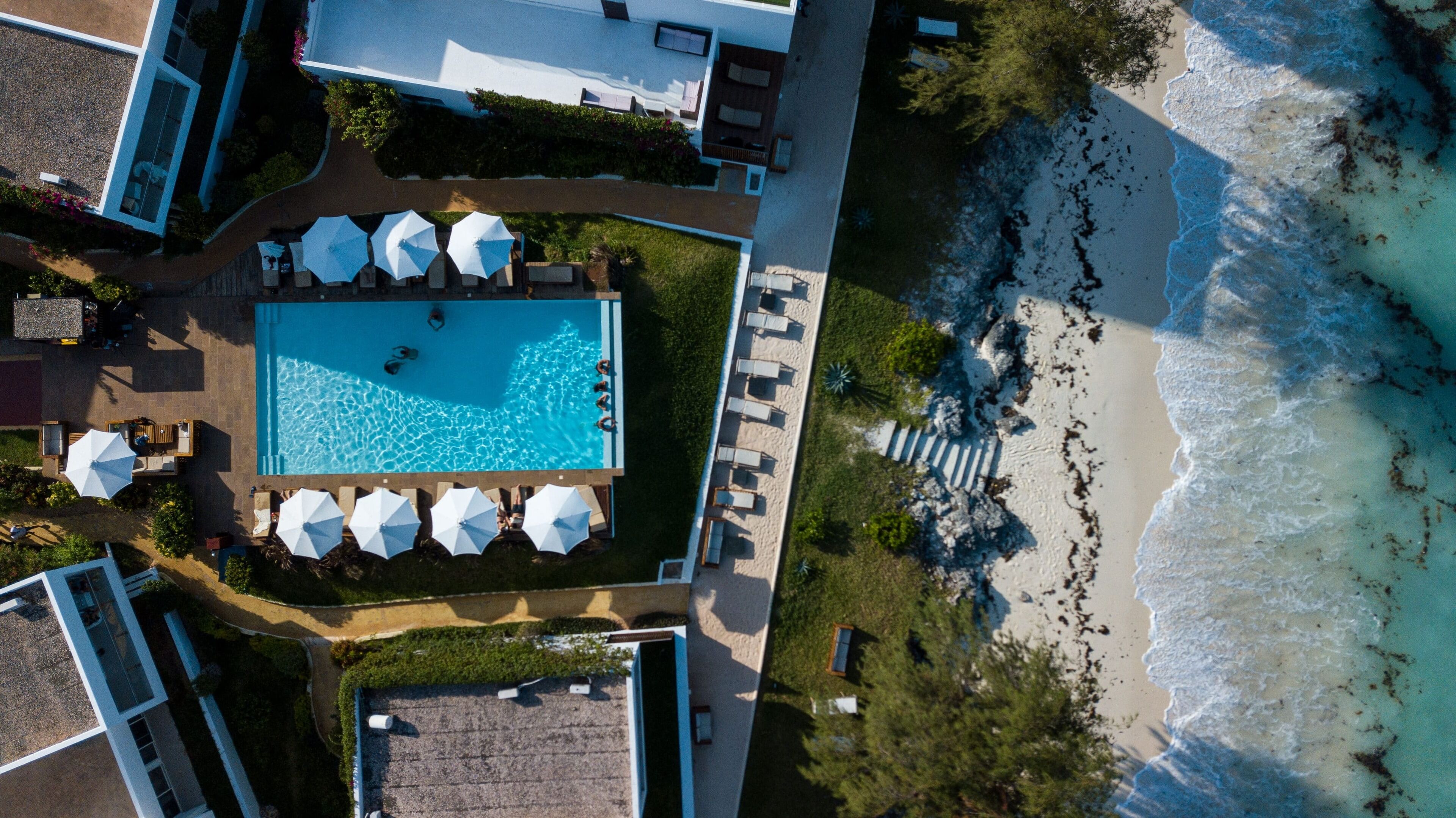 An aerial view of a resort with a swimming pool surrounded by lounge chairs and umbrellas, a beach, and a rocky shoreline.
