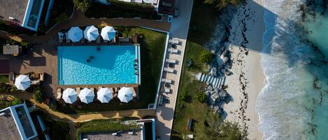 An aerial view of a resort with a swimming pool surrounded by lounge chairs and umbrellas, a beach, and a rocky shoreline.