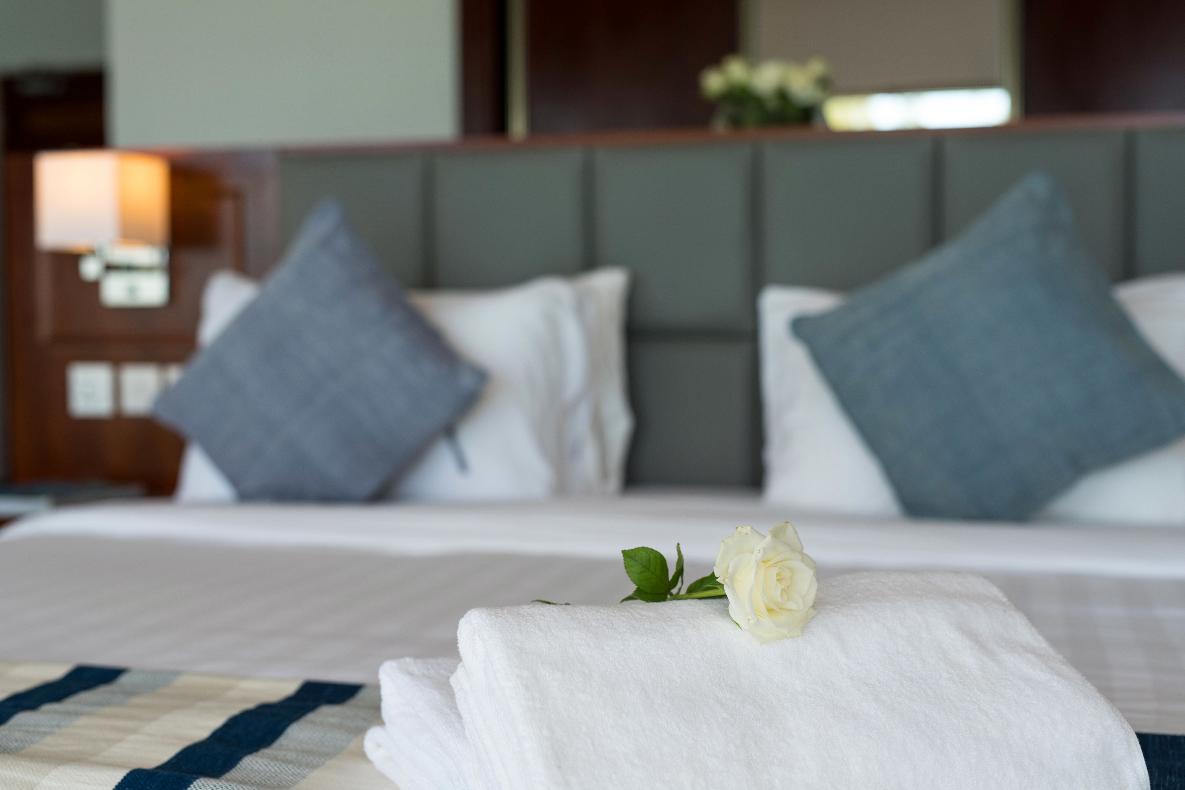 A neatly made bed with white linens, a single white rose, and two pillows, one grey and one white, in a hotel room.