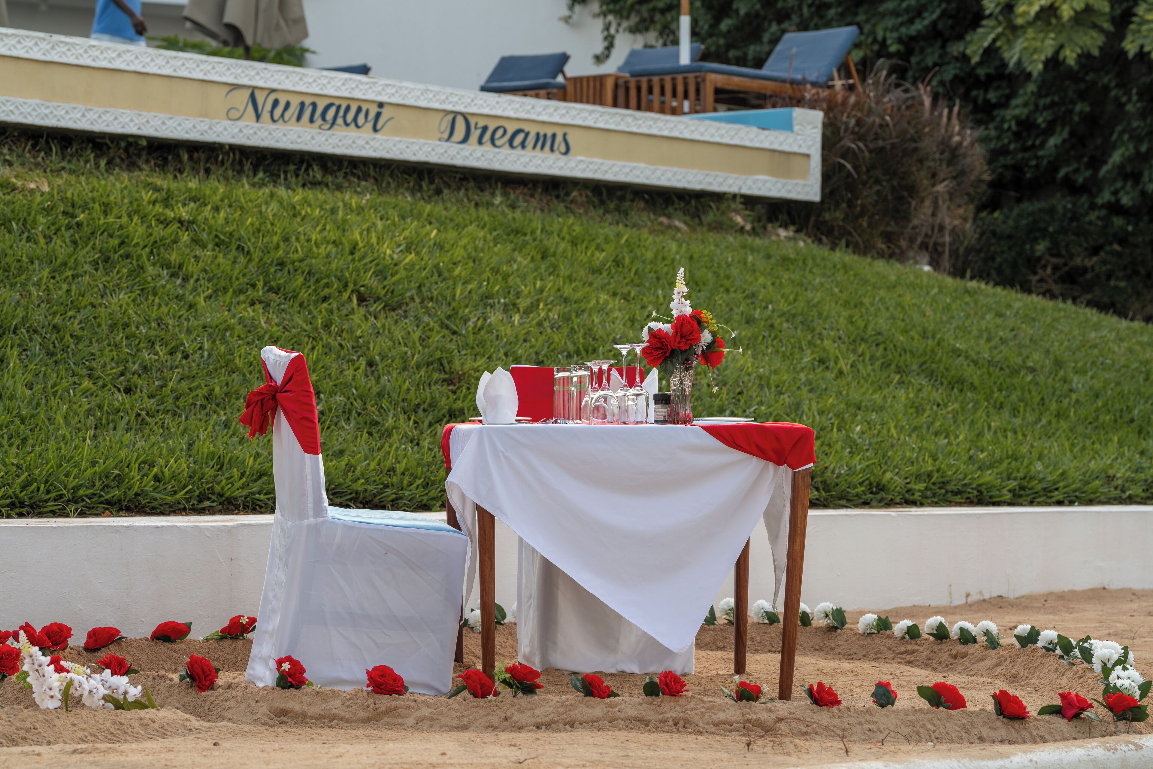 Une table dressée avec une nappe blanche et des touches de rouge, entourée de sable et de fleurs rouges et blanches.