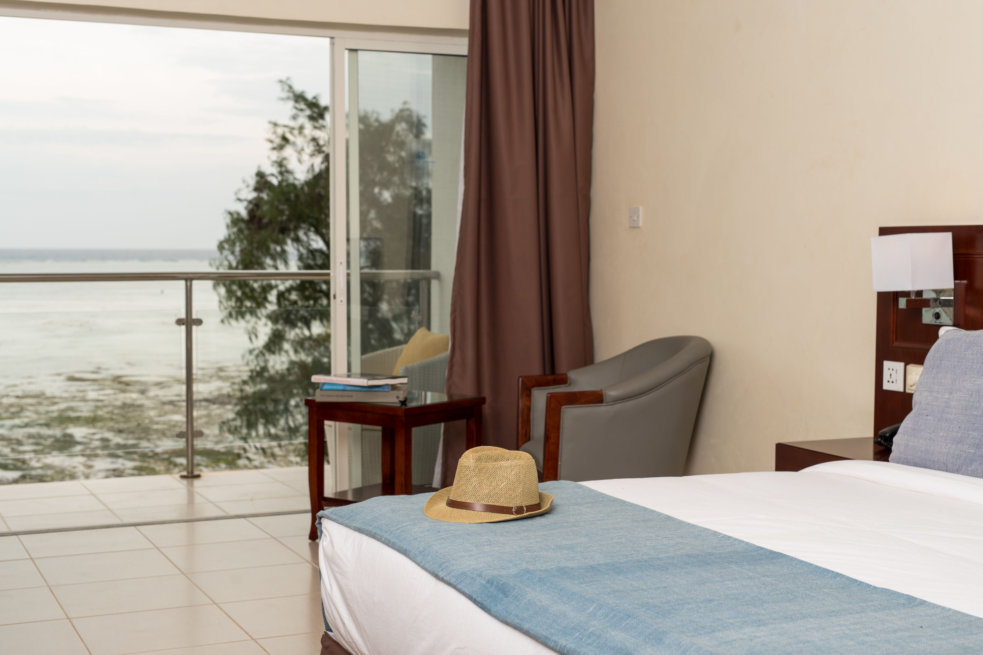 Ein Hotelzimmer mit Bett, Sessel, Nachttisch und Blick auf das Meer durch ein großes Fenster.