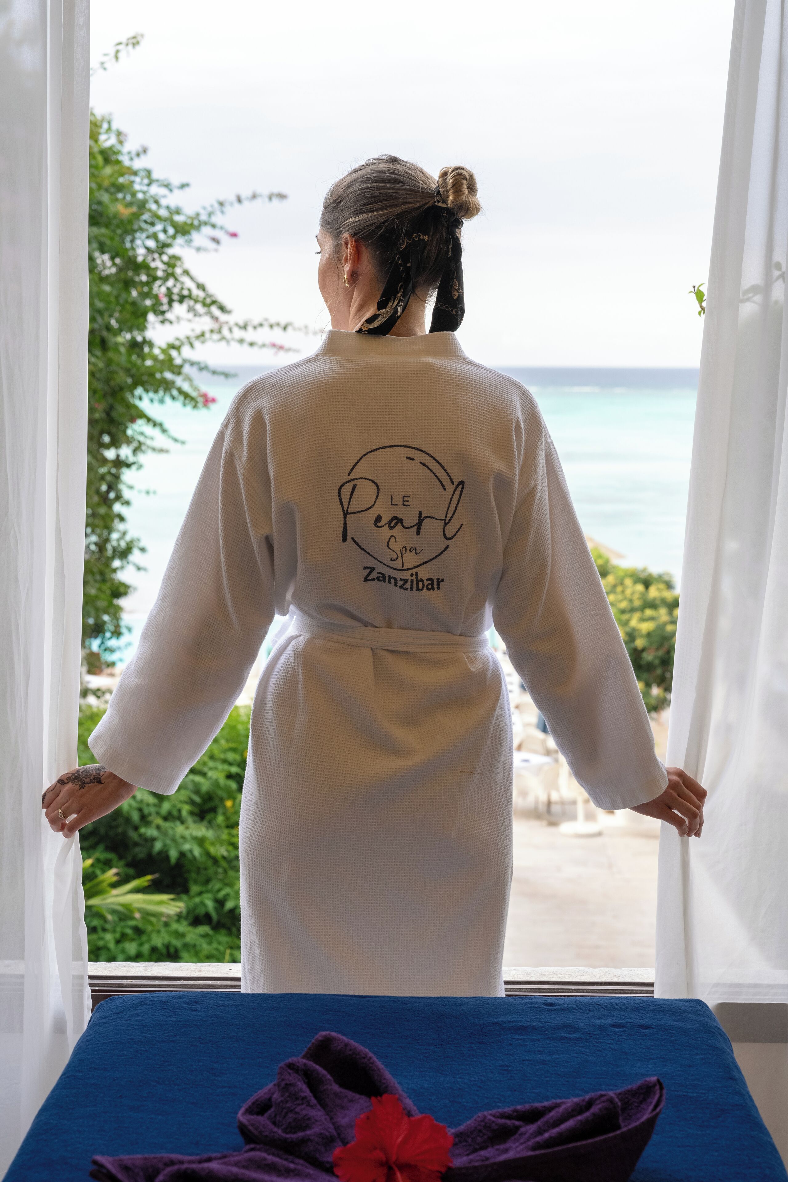 A person in a white robe standing by a window with a view of the sea.
