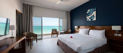 A hotel room with a bed, a desk, a chair, a TV, a ceiling fan, and a large window with a view of the ocean.