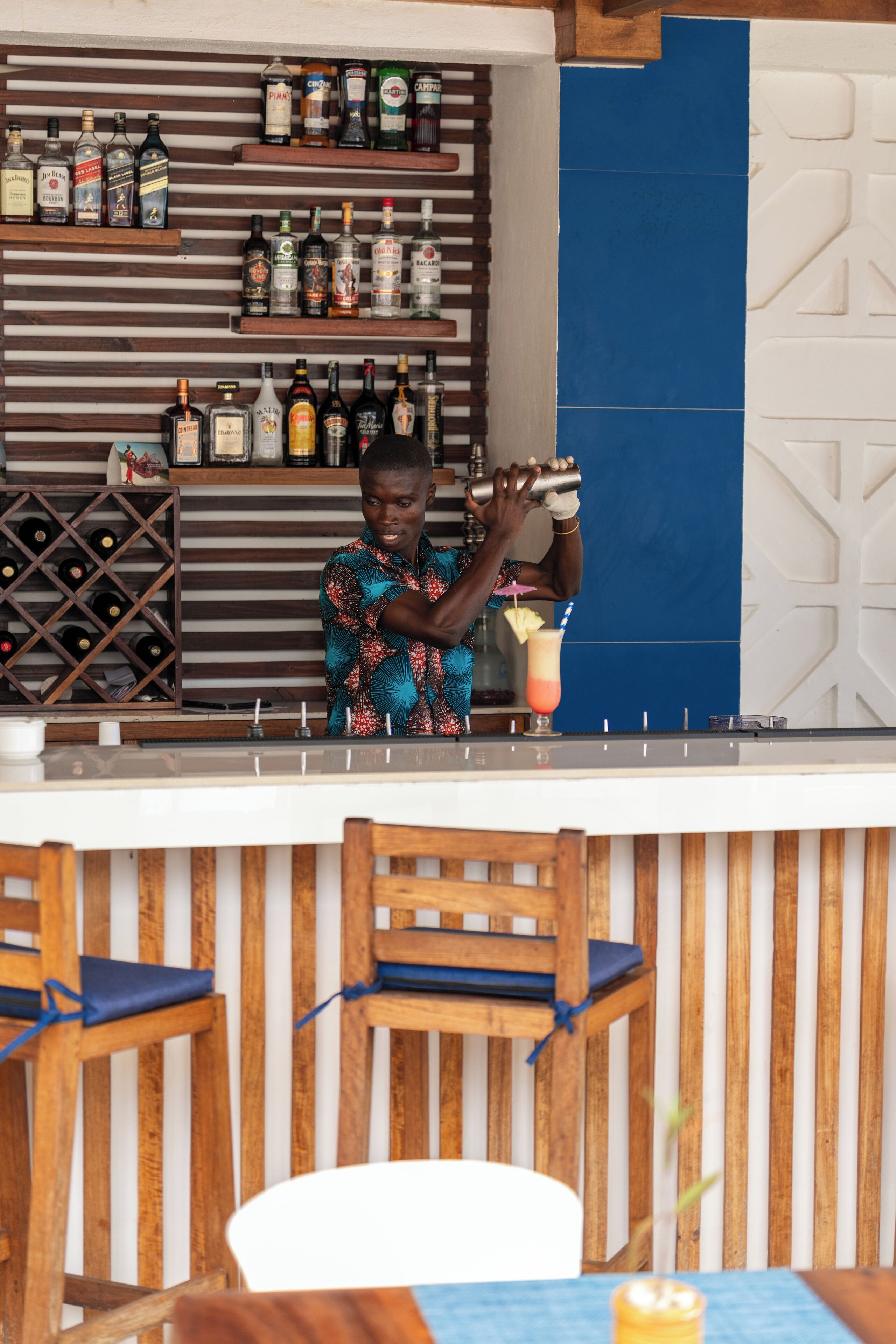 A bar area with wooden shelves displaying bottles, a bar counter, and wooden chairs with blue cushions.