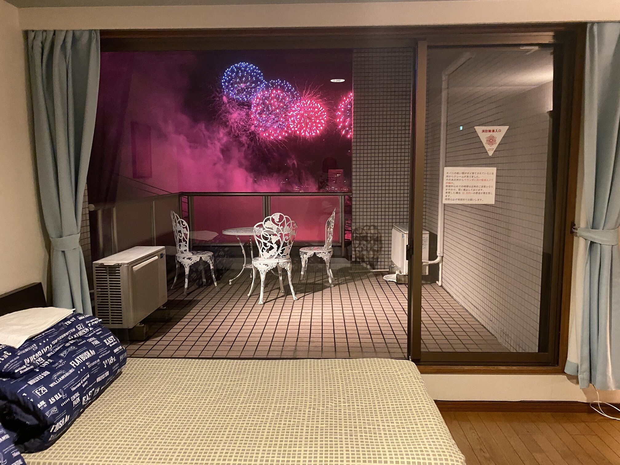A hotel room with a bed, a desk, and a chair. There is a view of fireworks in the background.