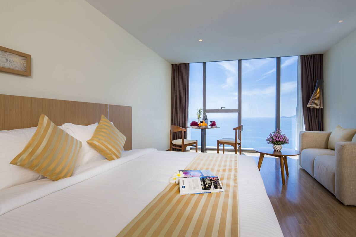 A hotel room with a large bed, a sofa, a small table with a vase of flowers, and a view of the sea.