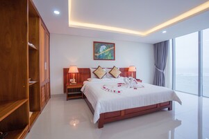 Un cuarto de hotel con una cama grande, cabecera de madera, lámparas de noche, un cuadro en la pared y vistas al mar a través de amplios ventanales.