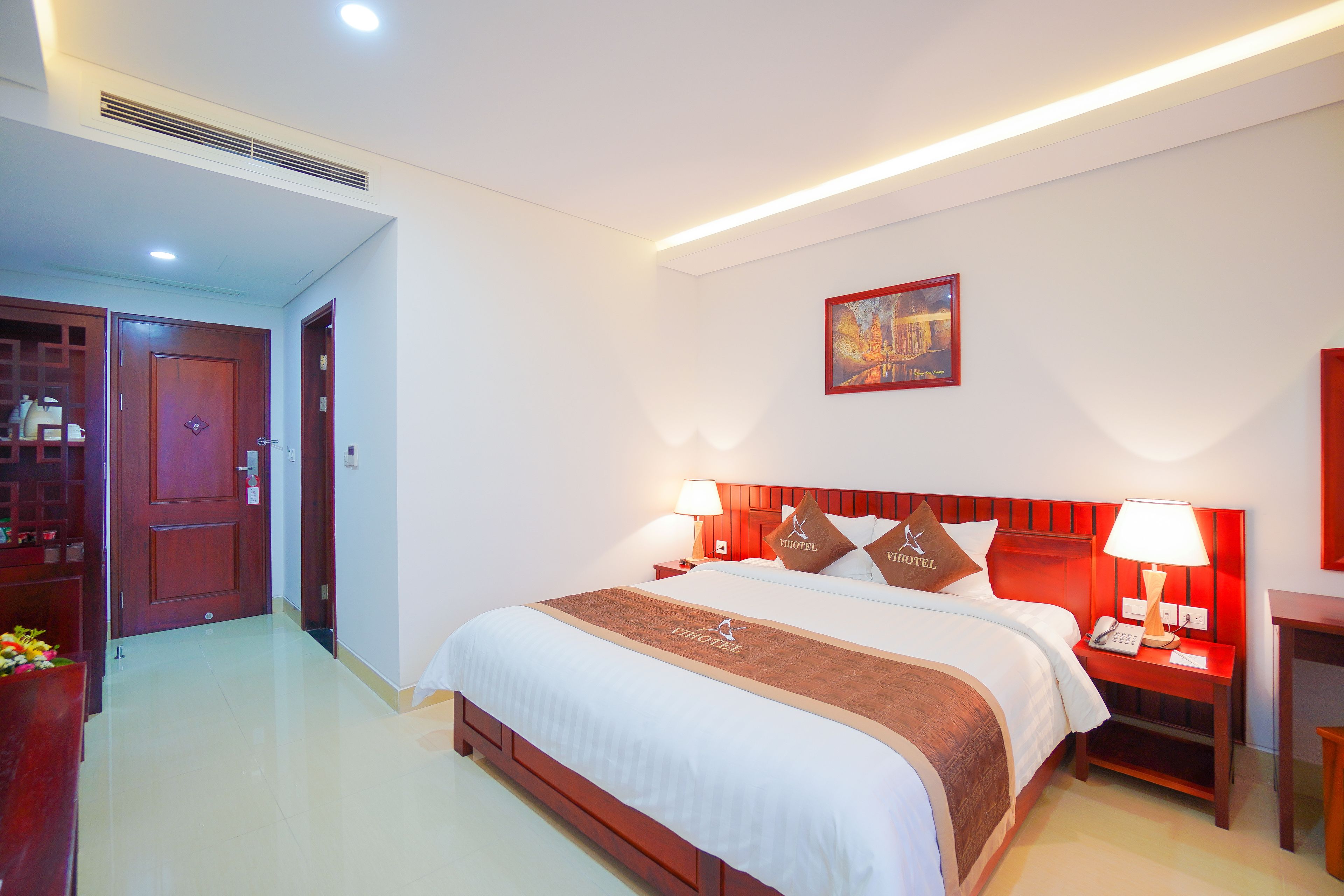 A hotel room with a large bed, two bedside tables with lamps, a wooden headboard, a painting on the wall, and a door leading to another room.