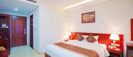 A hotel room with a large bed, two bedside tables with lamps, a wooden headboard, a painting on the wall, and a door leading to another room.
