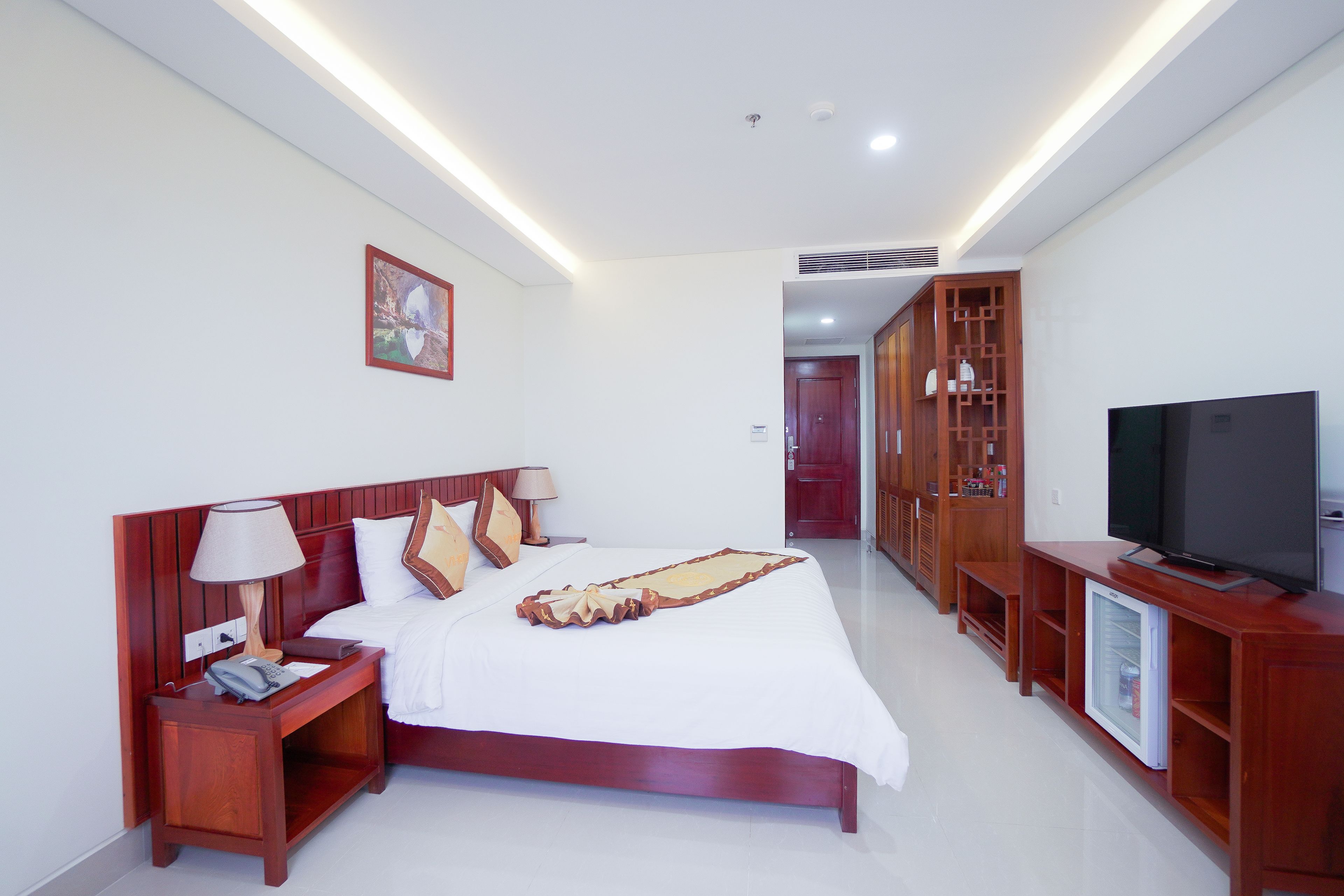 A hotel room with a wooden bed, bedside tables, a TV, and a mini-fridge.