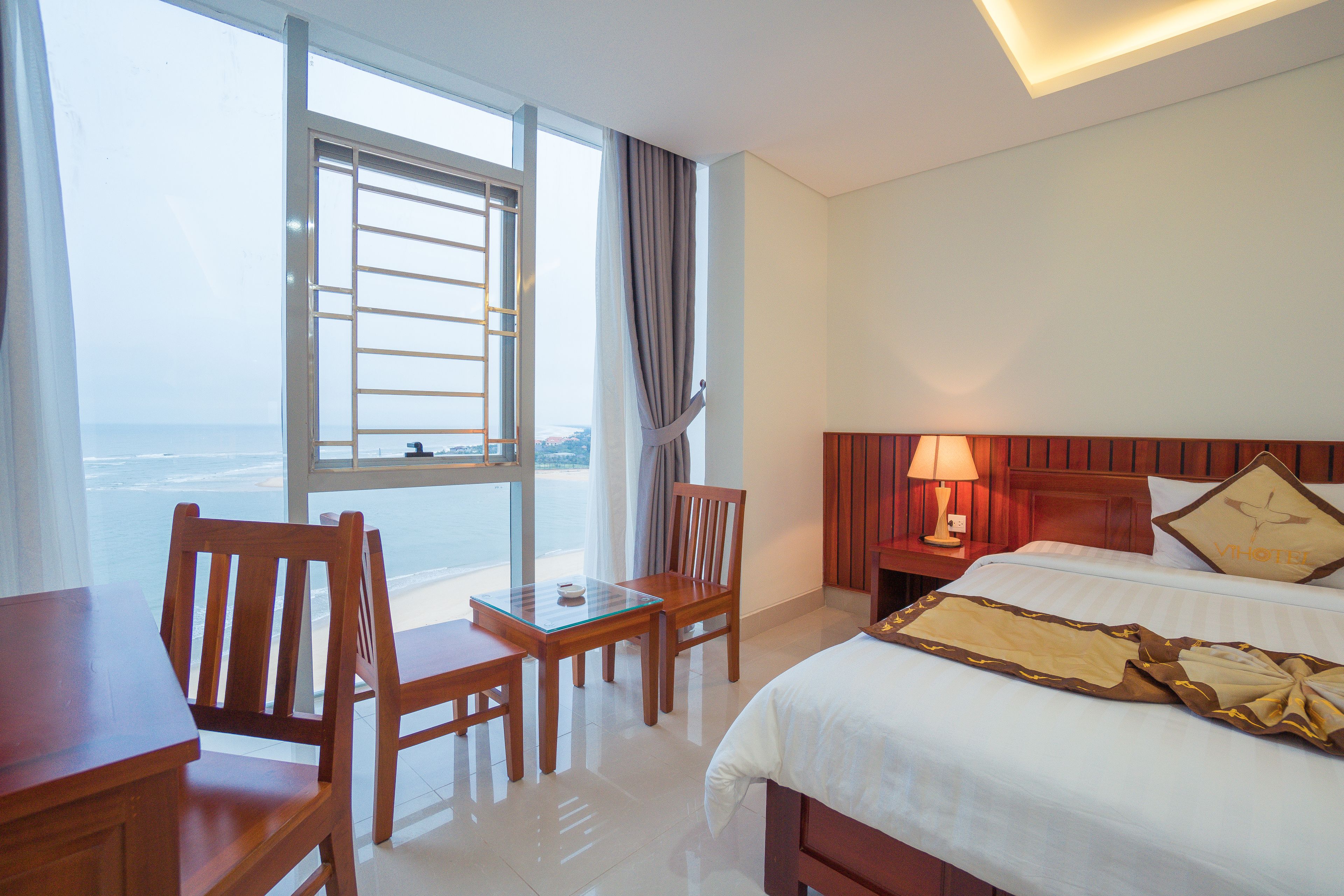 A hotel room with a bed, a desk, a chair, a nightstand, a lamp, and a view of the sea.