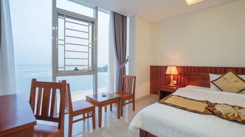 A hotel room with a bed, a desk, a chair, a nightstand, a lamp, and a view of the sea.