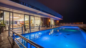 A swimming pool with a wooden deck and metal railings, surrounded by a building with large windows and indoor seating areas. - Vinh Hoang Hotel (Dong Hoi)