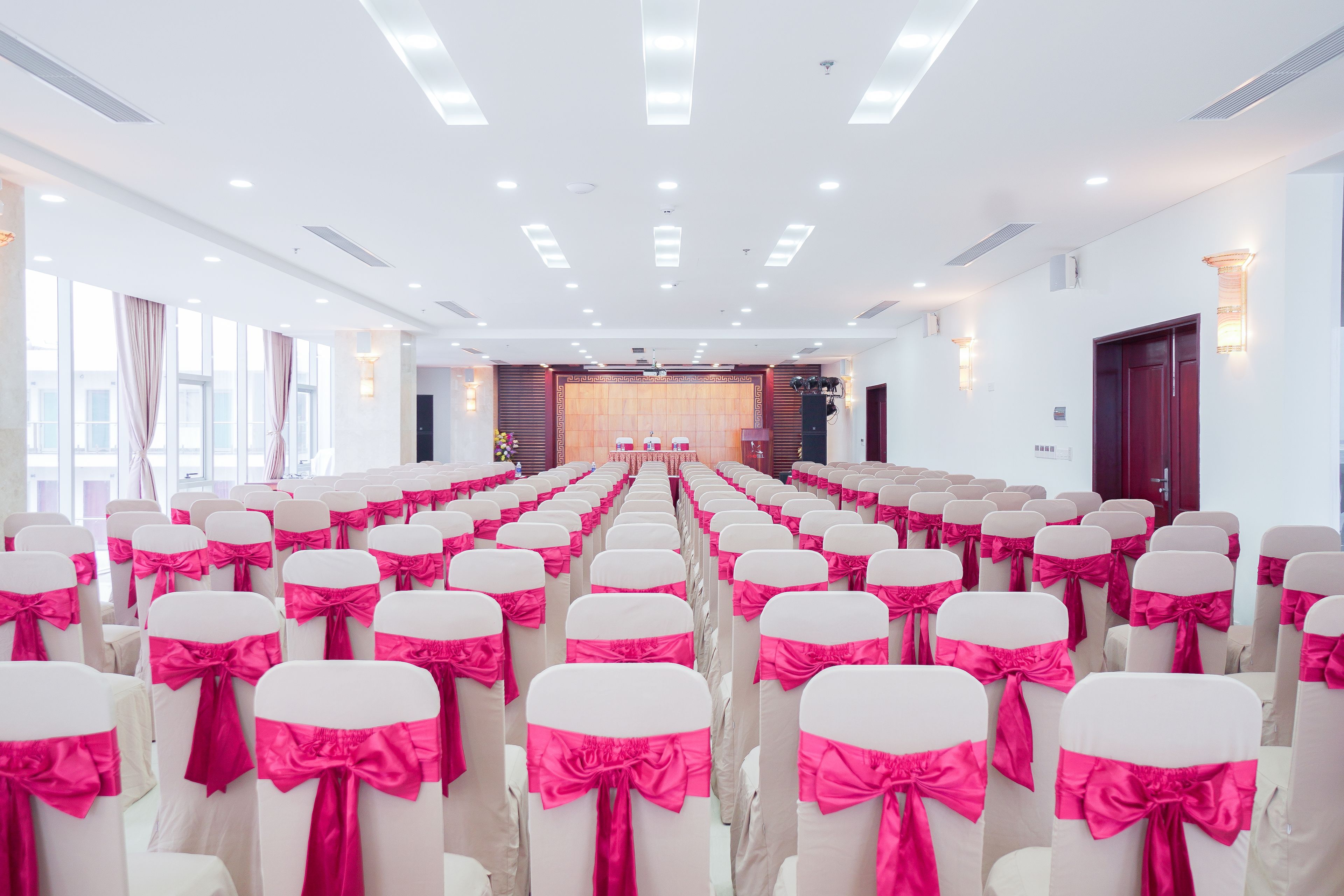A conference room with rows of chairs, each with a white cover and a pink bow. The room has a stage at the front, large windows on one side, and a door on the opposite wall.