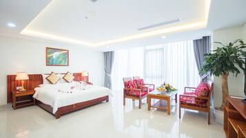 A hotel room with a large bed, two chairs, a small table, a plant, and a painting on the wall.