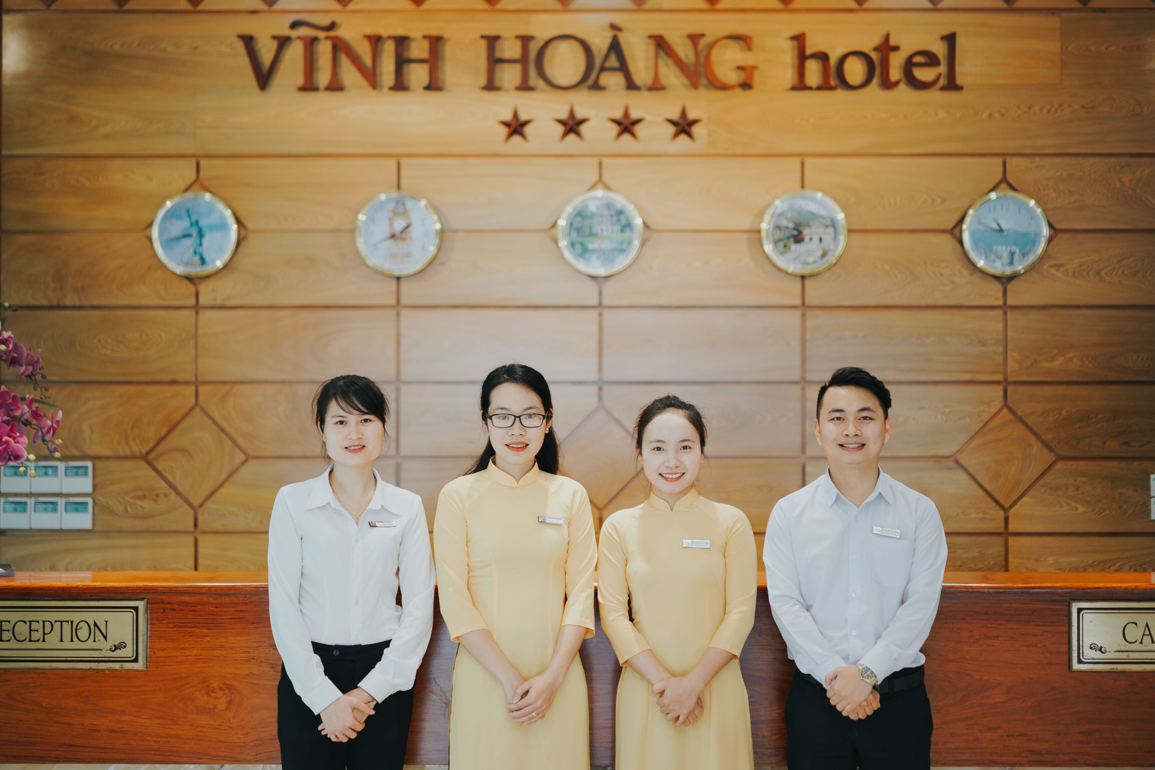 A reception desk at VINH HOANG hotel with a wooden panel design, clocks, and a floral arrangement.