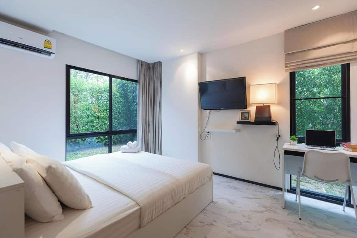 Ein modernes Hotelzimmer mit einem großen Bett, einem Schreibtisch mit Computer, einem Fernseher und großen Fenstern mit Blick auf Grünflächen.
