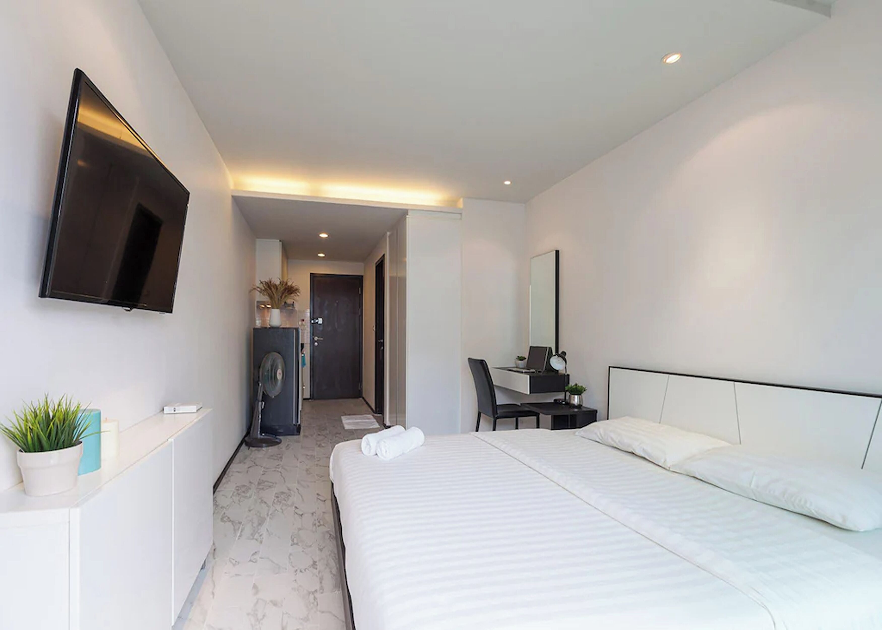 A modern hotel room with a large bed, a desk with a chair, a flat-screen TV mounted on the wall, and a small potted plant on the bedside table.